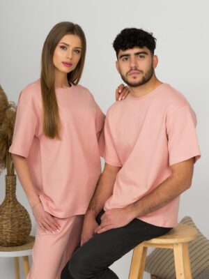 A young woman with blond hair wearing organic cotton pink t-shirt and pink straight leg trousers and a young man with black hair wearing organic cotton pink t-shirt and black trousers