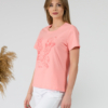 A young woman with blond hair wearing organic cotton peach color t-shirt with print and white trousers