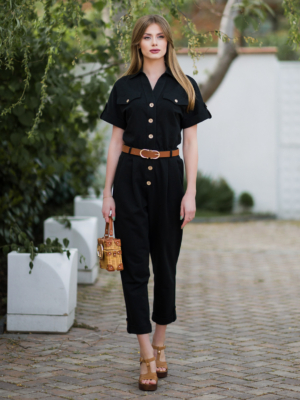 Young woman with blond hair wearing black organic cotton 7/8 length short sleeve jumpsuit with side pockets and coconut buttons