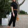 Young woman with blond hair wearing black organic cotton 7/8 length short sleeve jumpsuit with side pockets and coconut buttons