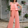 A woman stands outdoors on a stone path, wearing a peach color two-piece outfit and white sandals. She is near a covered seating area with floral cushions and greenery in the background.