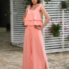 A woman stands outdoors on a stone path, wearing a peach color two-piece outfit and white sandals. She is near a covered seating area with floral cushions and greenery in the background.