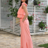 A woman stands outdoors on a stone path, wearing a peach color two-piece outfit and white sandals. She is near a covered seating area with floral cushions and greenery in the background.