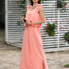 A woman stands outdoors on a stone path, wearing a peach color two-piece outfit and white sandals. She is near a covered seating area with floral cushions and greenery in the background.