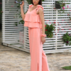 A woman stands outdoors on a stone path, wearing a peach color two-piece outfit and white sandals. She is near a covered seating area with floral cushions and greenery in the background.