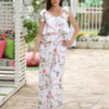 A woman stands outdoors on a stone path, wearing a white floral two-piece outfit and white sandals. She is near a covered seating area with floral cushions and greenery in the background.