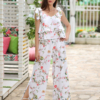 A woman stands outdoors on a stone path, wearing a white floral two-piece outfit and white sandals. She is near a covered seating area with floral cushions and greenery in the background.