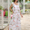 A woman stands outdoors on a stone path, wearing a white floral two-piece outfit and white sandals. She is near a covered seating area with floral cushions and greenery in the background.