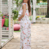 A woman stands outdoors on a stone path, wearing a white floral two-piece outfit and white sandals. She is near a covered seating area with floral cushions and greenery in the background.