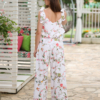 A woman stands outdoors on a stone path, wearing a white floral two-piece outfit and white sandals. She is near a covered seating area with floral cushions and greenery in the background.
