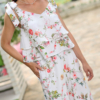 A woman stands outdoors on a stone path, wearing a white floral two-piece outfit and white sandals. She is near a covered seating area with floral cushions and greenery in the background.