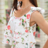 A woman stands outdoors on a stone path, wearing a white floral two-piece outfit and white sandals. She is near a covered seating area with floral cushions and greenery in the background.