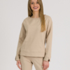 woman with blond hair wearing beige organic cotton long sleeve off shoulder boat neck blouse and beige organic cotton long tapered leg pants