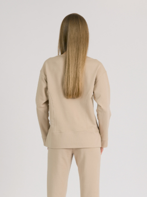woman with blond hair wearing beige organic cotton long sleeve off shoulder boat neck blouse and beige organic cotton long tapered leg pants