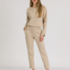 woman with blond hair wearing beige organic cotton long sleeve off shoulder boat neck blouse and beige organic cotton long tapered leg pants
