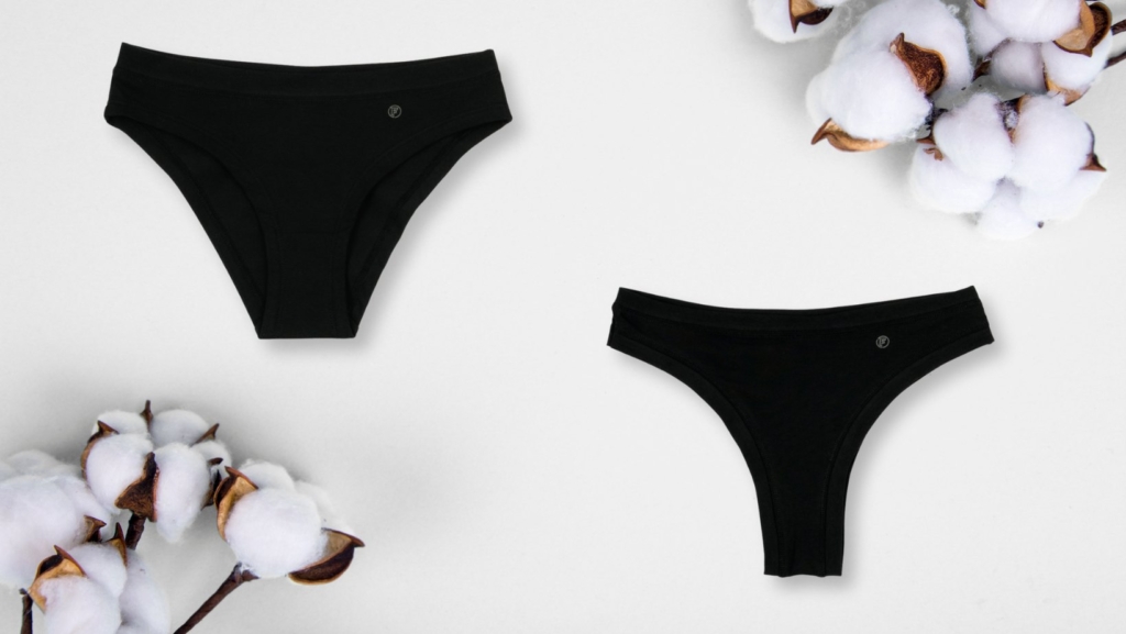 Two pairs of black organic cotton underwear are placed on a light surface, surrounded by cotton branches.