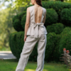 A dark-haired woman wearing a beige elegant top with a belt at the waist, a V-neckline and an open back, and beige pants made of 100% linen.