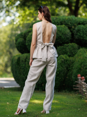 A dark-haired woman wearing a beige elegant top with a belt at the waist, a V-neckline and an open back, and beige pants made of 100% linen.