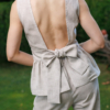 A dark-haired woman wearing a beige elegant top with a belt at the waist, a V-neckline and an open back, and beige pants made of 100% linen.