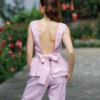 A dark-haired woman wearing a pink elegant top with a belt at the waist, a V-neckline and an open back, and pink pants made of 100% linen.