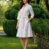 A woman stands on grass in front of trimmed green bushes, wearing a beige and white striped button-down dress with a belt and white heels.