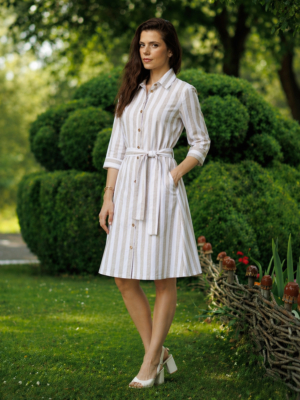 A woman stands on grass in front of trimmed green bushes, wearing a beige and white striped button-down dress with a belt and white heels.