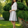 A woman stands on grass in front of trimmed green bushes, wearing a beige and white striped button-down dress with a belt and white heels.