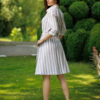 A woman stands on grass in front of trimmed green bushes, wearing a beige and white striped button-down dress with a belt and white heels.