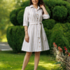 A woman stands on grass in front of trimmed green bushes, wearing a beige and white striped button-down dress with a belt and white heels.