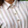 A woman stands on grass in front of trimmed green bushes, wearing a beige and white striped button-down dress with a belt and white heels.