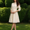 A woman stands on grass in front of trimmed green bushes, wearing a yellow and white striped button-down dress with a belt and white heels.