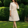 A woman stands on grass in front of trimmed green bushes, wearing a yellow and white striped button-down dress with a belt and white heels.