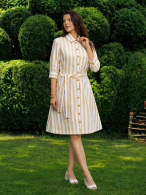 A woman stands on grass in front of trimmed green bushes, wearing a yellow and white striped button-down dress with a belt and white heels.