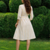 A woman stands on grass in front of trimmed green bushes, wearing a yellow and white striped button-down dress with a belt and white heels.