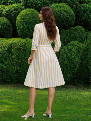 A woman stands on grass in front of trimmed green bushes, wearing a yellow and white striped button-down dress with a belt and white heels.