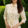 A woman stands on grass in front of trimmed green bushes, wearing a yellow and white striped button-down dress with a belt and white heels.